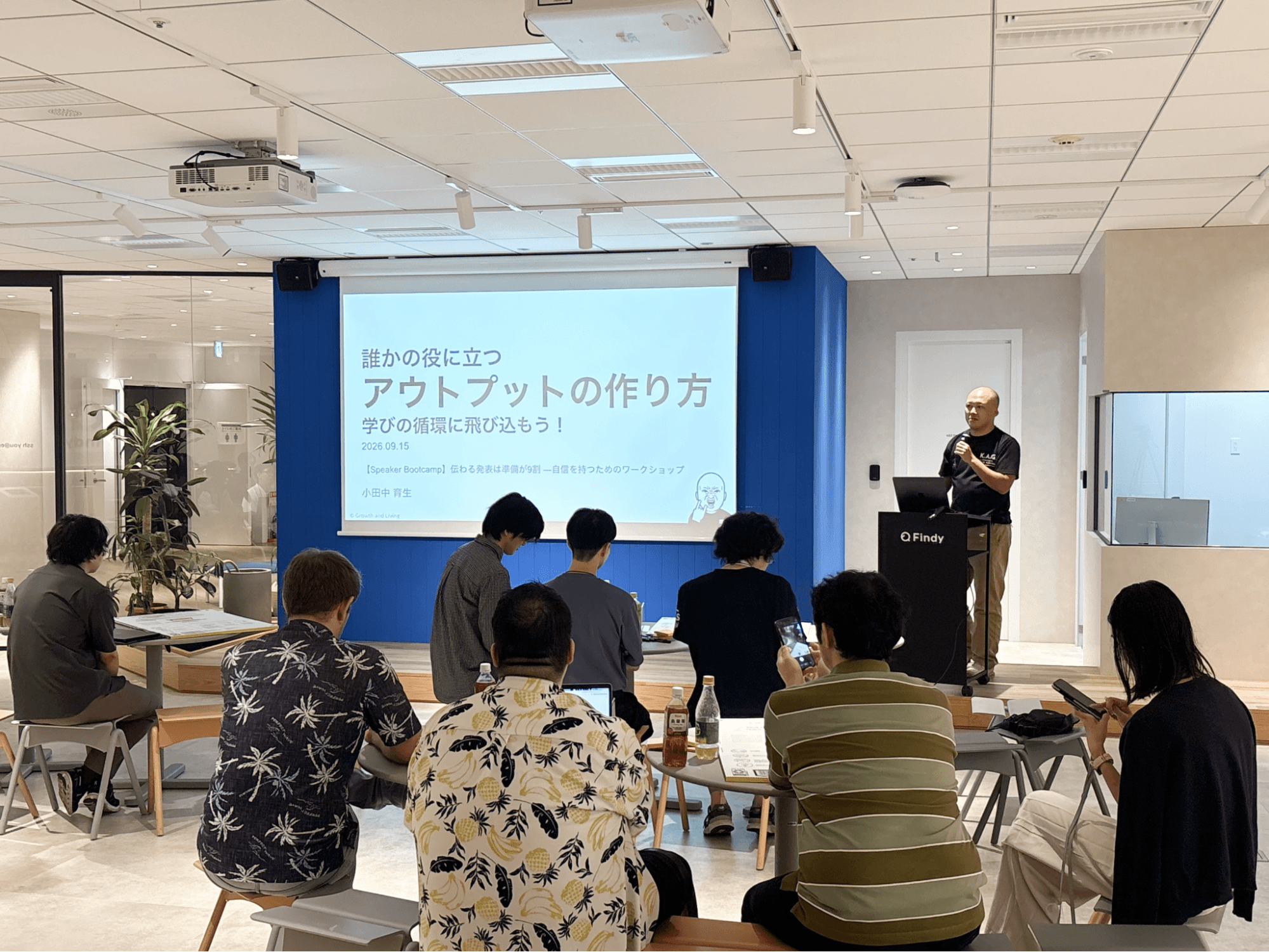 「誰かの役に立つアウトプットの作り方 学びの循環に飛び込もう!」のタイトルスライドを背に、演台でマイクを持って話す小田中育生さん。手前の丸テーブルでは参加者が着席して聞いている